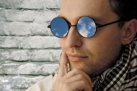 Thoughtful young man wearing glasses with the reflection of blue sky (against the background of a...