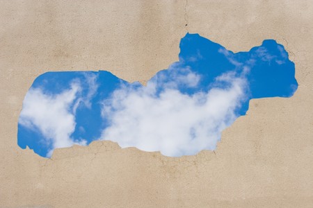 Gray wall with peeling plaster (with blue sky and clouds showing through the hole)の写真素材