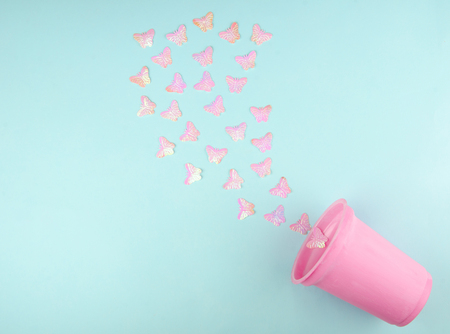 Beautiful pink artificial butterflies flying out of a pink plastic glass (on a blue paper background), minimal concept, flat lay with copy space on the leftの写真素材