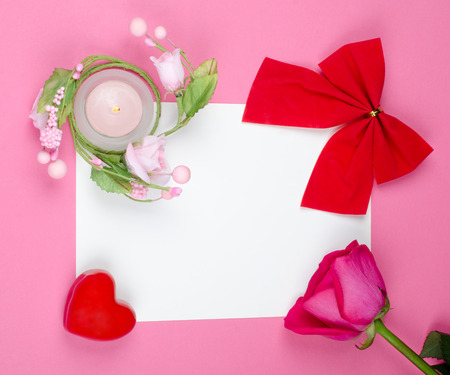 Bright red bow, pink rose, candle and red heart on a blank white greeting card against a bright pink background (flat lay, top view)の写真素材