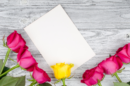 Beautiful pink roses and a single yellow rose on a blank white greeting card against a worn wooden background (flat lay, top view)の写真素材