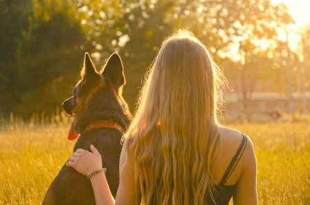 Blurred silhouette of a blonde girl embracing a German shepherd, sitting in the grass in the golden rays of the sun and looking at the sunset (selective focus on the girl), retro style tonedの写真素材