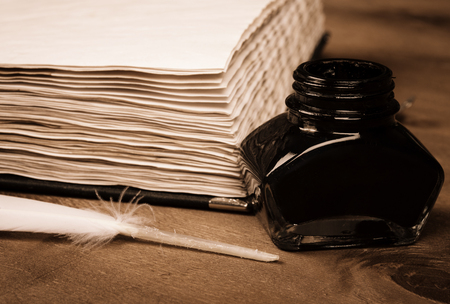 Blank old diary, an inkwell and a feather as a quill pen on a worn wooden background (selective focus on the inkwell and feather), retro style in sepiaの写真素材