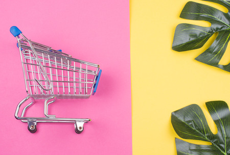 Shopping cart and monstera leaves on the bright pink and yellow background (summer sale concept, top view), selective focus on the shopping cartの写真素材