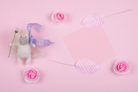 Tiny unicorn toy made of wool, handmade rose buds, handmade pink leaves and a pink paper card on a pastel pink backgroundの写真素材