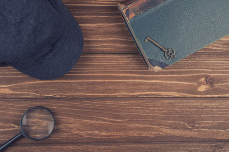 Cap of a detective, a magnifying glass and an old book with a key on the book against a worn wooden background (top view, retro style) as the detective or crime story conceptの写真素材