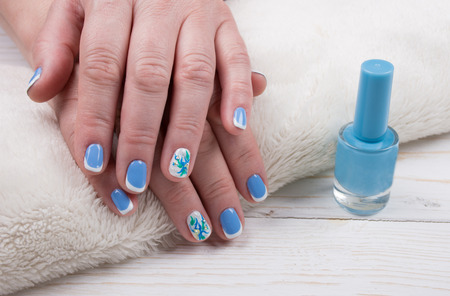 Female hands with manicured nails on a white towel and a bottle of nail polish of blue color against the white wooden background (manicure concept)の写真素材