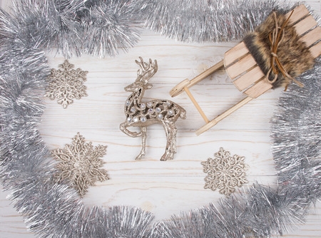 Toy reindeer, wooden Christmas sleigh, silver tinsel and snowflakes on a white wooden background (top view)の写真素材
