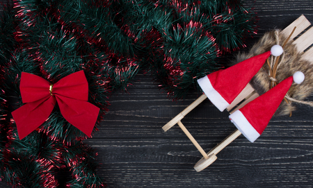 Two Santa hats on a toy Christmas sleigh, Christmas bow and green and red tinsel on a black wooden background as a New Year or Christmas background (top view, copy space for your text)の写真素材
