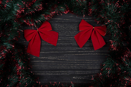 Two bright red Christmas bows and green and red tinsel forming a frame on a black wooden background as a New Year or Christmas background (top view, copy space for your text)の写真素材