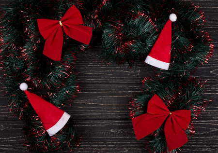 Santa hats, red Christmas bows and green and red tinsel forming a frame on a black wooden background as a New Year or Christmas background (top view, copy space in the center for your text)の写真素材
