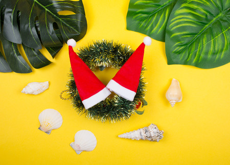 Two small Santa hats on a Christmas wreath among tropical leaves and seashells on the bright yellow background as the tropical Christmas conceptの写真素材