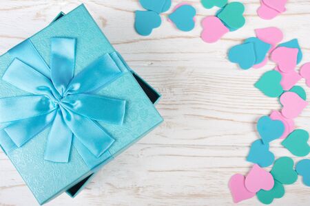 Blue gift box and a pile of paper hearts of pastel colors on the white wooden background (flat lay,...
