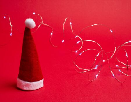 Santa hat and a garland with Christmas lights on the bright red background as the Christmas concept (selective focus on the Santa hat)の写真素材