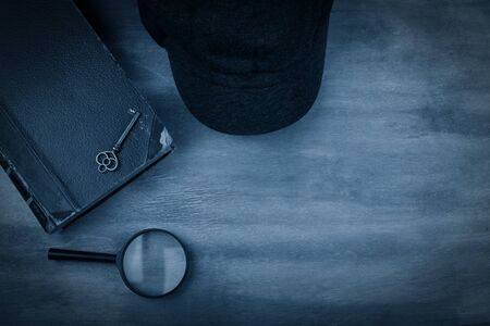 Old book, a key, a detective cap and a magnifying glass against a worn wooden background with dark edges toned in the classic blue color (top view) as the crime story conceptの写真素材