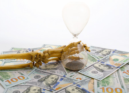 skeleton fingers holding sand-glass placed on dollars. concept of time - money and death.