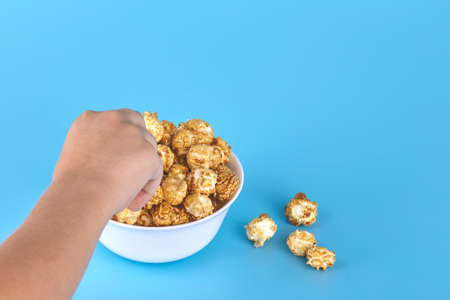 The child takes sweet popcorn from a white plate. Blue background, free space. The concept of...