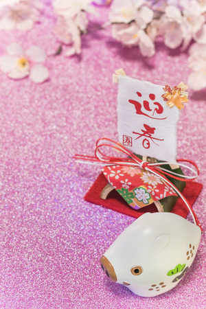 Pink glitter background with sakura cherry blossoms for japanese New Year's Cards with cute animal figurine of boar or pig and rice paper on bamboo stand with handwriting ideograms Geishun which means Welcome Spring.の写真素材