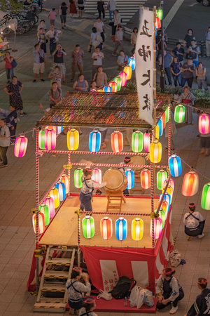 View of the square in front of the Nippori train station decorated for the Obon festival with a yagura tower illuminated with paper lanterns where a girl in traditional costume is playing taiko drum in the summer night of Arakawa district in Tokyo.のeditorial素材