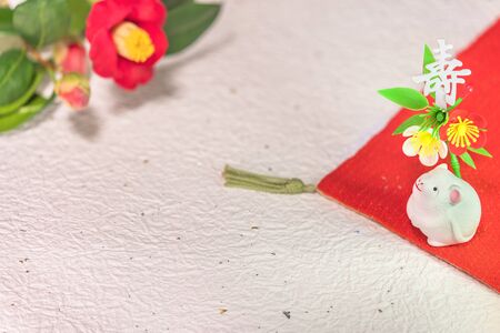 A Japanese greeting card with a Tsubaki flower also called the winter rose and a cute rat figurine for the 2020 year of the mouse on a red cloth decorated with the Japanese Kotobuki ideograph which means longevity on a washi crumpled paper with golden and silver fragments.の写真素材