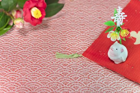 A Japanese greeting card with a Tsubaki flower also called the winter rose and a cute rat figurine for the 2020 year of the mouse on a red cloth decorated with the Japanese Kotobuki ideograph which means longevity.の写真素材