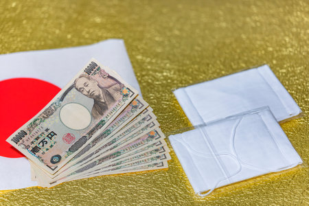 tokyo, japan - may 20 2020: Cloth masks and 100,000 yen in cash sent by the Japanese govt of Abe Shinzo to fight against coronavirus (COVID-19) and unemployment on a Japan flag and golden background.のeditorial素材