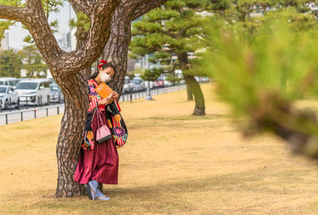 Tokyo, Japan - March 01 2020: Japanese woman dressed in a hakama kimono and wearing a facial mask reading a Tokyo travel guide book leaned against the trunk of pine tree in the Kokyo Gaien National Garden of Tokyo Imperial Palaceのeditorial素材