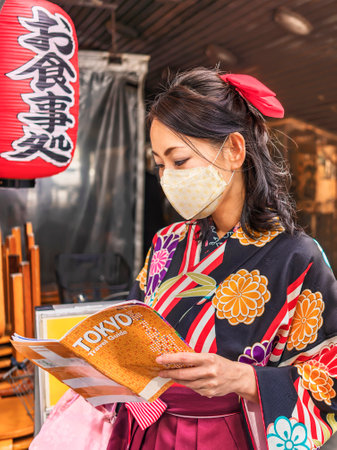 Tokyo, Japan - March 01 2020: Japanese woman in hakama kimono wearing a cloth mask and reading a Tourist guide book of Tokyo city at Yuraku Concourse underpass decorated with a red paper lantern where is inscribed the word meal.のeditorial素材