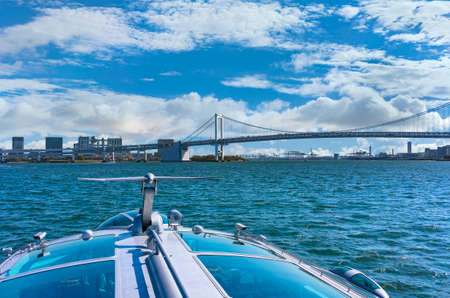 Aerodynamic fin on the rooftop of a waterbus sailing to double-layered suspension Rainbow Bridge with the bay of Odaiba and malls buildings in the background.の写真素材