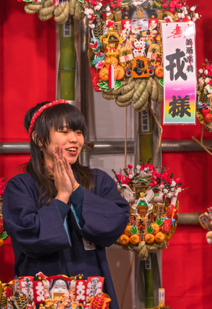 asakusa, japan - november 08 2019: Young female merchant in a stall of the Rakes Fair or Tori-no-Ichi Fair doing a tejime hand clapping after selling to customers an auspicious rake in Ootori shrine.のeditorial素材