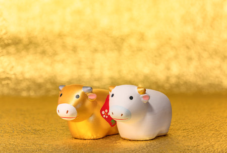 Greeting card with a close-up on two cute hand made figurines of the zodiac animal of two cows hand painted in gold and white for the year of the ox on an uneven textured golden background.の写真素材