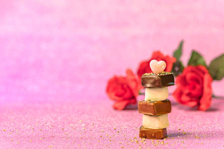 Valentine or white day greeting card depicting a column of stacked chocolates topped with a heart shaped object against a background of red roses flowers on a glittering pink paper with gold nuggets.の写真素材