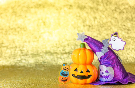 Concept photography depicting an halloween jack o lantern pumpkin head and a purple witches hat with funny ghost smiling and cardboard cutout bats against a shiny golden background.の写真素材