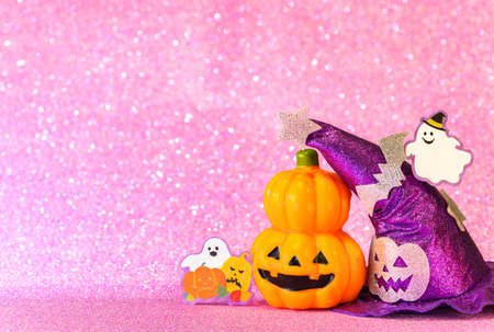 Concept photography depicting an halloween jack o lantern pumpkin head and a purple witches hat with funny ghost smiling and cardboard cutout bats against a glittering pink background.の写真素材