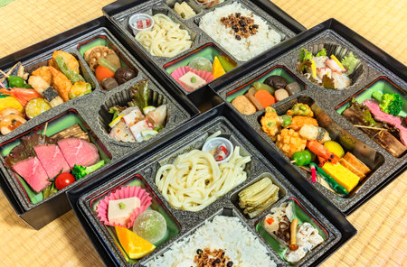 Deluxe Japanese Makunouchi bento boxes with rice,udon noodles, assortment of tsukemono preserved and nimono simmered vegetables, roast beef slices, sashimi, tofu, wagashi confectioneries on a tatami.の写真素材