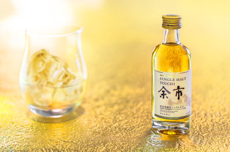 Hokkaido, japan - April 05 2023: A small bottle miniature of single malt Yoichi from the Japanese maker Nikka Whiskey displayed on a golden surface with an on the rock glass in a shallow depth of field.のeditorial素材
