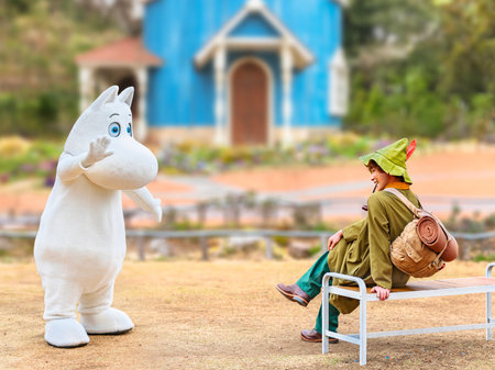 japan, saitama - mar 26 2023: Moomintroll costume waving hand to his smiling friend Snufkin wearing a green hat carrying a backpack with a camp mattress smoking a pipe on a bench in Moominvalley park.のeditorial素材