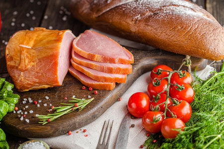 Smoked ham on a cutting board with herbs and tomatoes on a wooden backgroundの写真素材