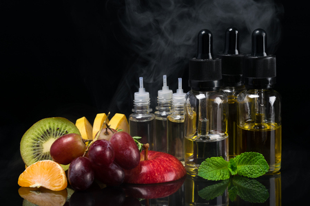 Fruit and flavors in bottles for an electronic cigarette, concept on a black background with a steamの写真素材
