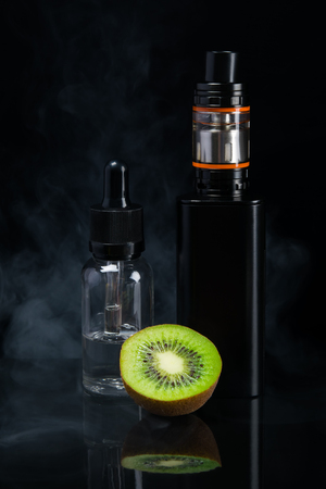 Liquid with a taste of kiwi next to an electronic cigarette, on a black background in smokeの写真素材