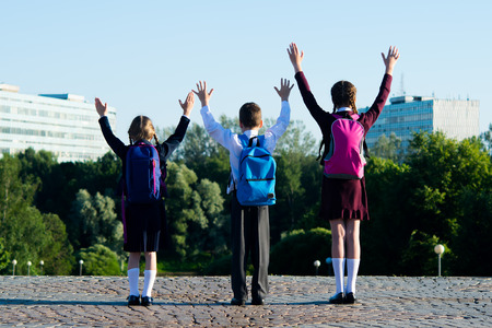 Three schoolchildren amicably walking in the park, and raise their hands upwardsの写真素材