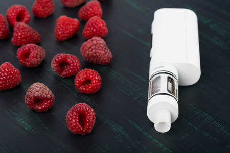 white electronic cigarette, on a black painted board, next to a red raspberryの写真素材