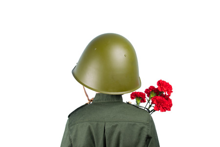 a child in military uniform and a helmet stands with his back, holding a carnation, in front of a white backgroundの写真素材