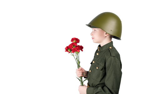 a boy in a helmet wearing a military uniform stands facing sideways and holding red flowers, isolated on whiteの写真素材