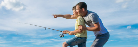 against the blue sky, the son holds a fishing rod, and the father shows in the distanceの写真素材