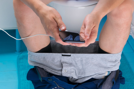 a man sitting on the toilet, with his pants down and holding a mobile phone that is charging