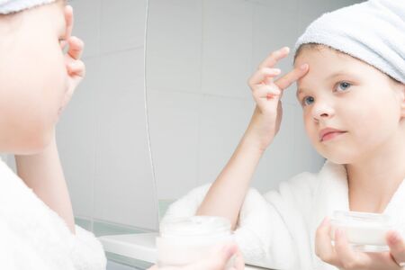 girl in a white coat with a towel on her head with fingers applies cream to her faceの写真素材