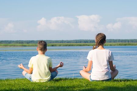 boy and girl sit on the green grass and do yoga, rear view, against the backdrop of a beautiful landscapeの写真素材