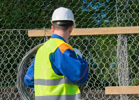 worker in a yellow vest and a white helmet, carries a roll of wire in the shoulder, rear viewの写真素材