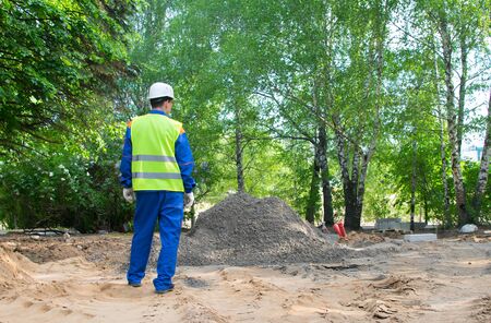 a worker in a blue uniform and a yellow vest, stands with his back in front of a pile of soil to...